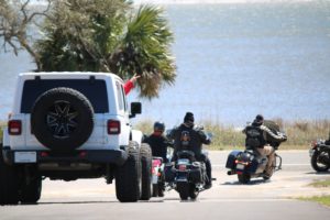A jeep and several motorcycles pull out of driveway with the bay in the background